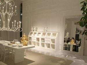 foto interna dello stand con credenza laccata bianca con gioco di pieni e vuoti tra cassetti in legno ed ante in vetro, sulla sinistra tavolo da pianto con grande lampadario scenografico sempre bianco